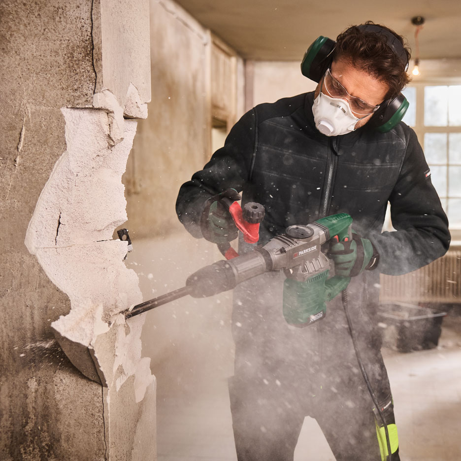 Man with demolition hammer, mask, and ear protection demolishing a wall.