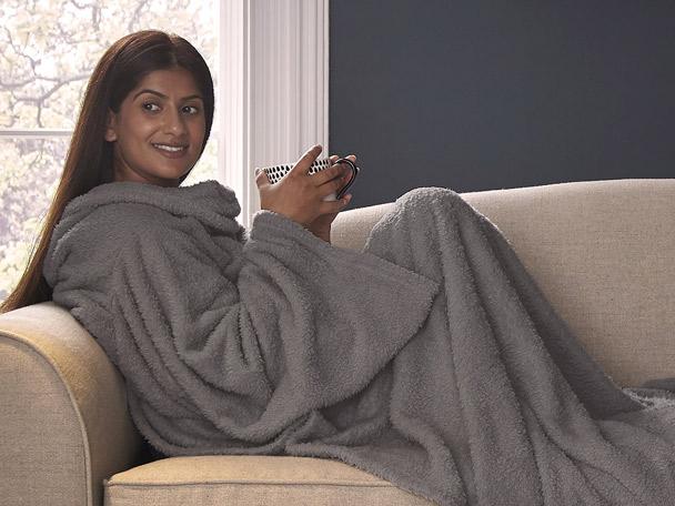 Woman on a sofa, wrapped in a grey hooded blanket, holding a mug.