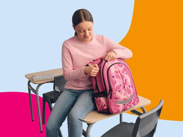 Girl in a classroom setting, sitting on a desk and opening a pink floral backpack.