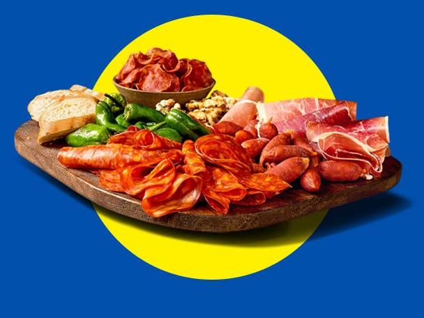 A wooden board with various cured meats, green peppers, bread, and nuts on a blue and yellow background.