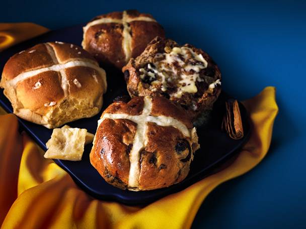 Hot cross buns on a dark plate with a yellow cloth and cinnamon sticks.