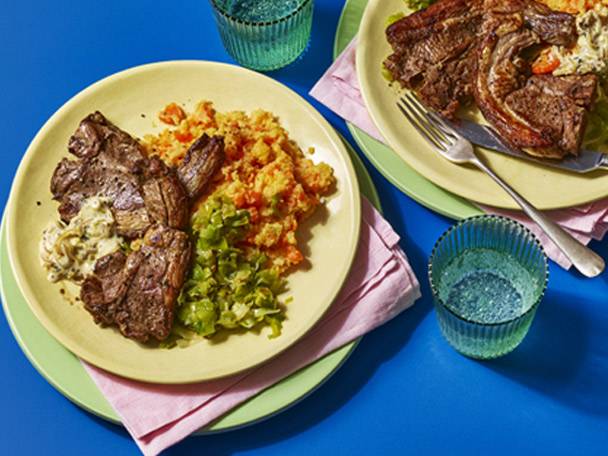 Two plates of lamb chops with mashed vegetables and a side of green vegetables.