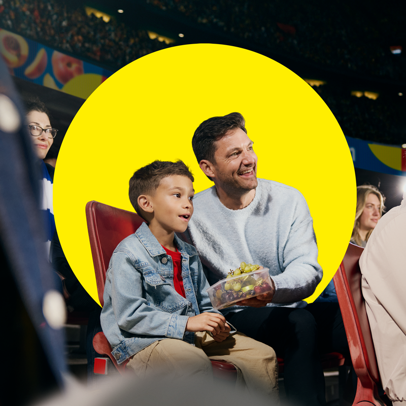 Father and son enjoy fruit in a container during a sporting event.