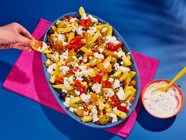 A hand dipping a chip into a large platter of loaded fries with feta, tomatoes, and olives.
