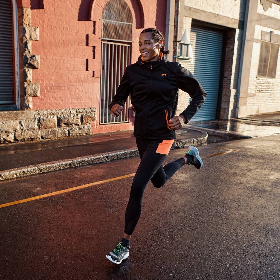 Man in black running jacket and leggings with orange accents, running on a wet street.